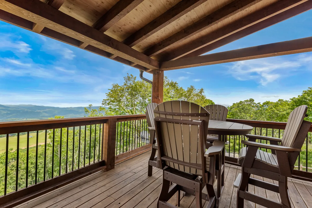 A serene wooden deck furnished with comfortable outdoor seating overlooks a breathtaking panoramic view of the surrounding mountains and forest. The setting is tranquil, with space for couples to relax, entertain, or simply soak in nature's beauty. Soft evening light suggests it’s perfect for sunset viewing or stargazing.