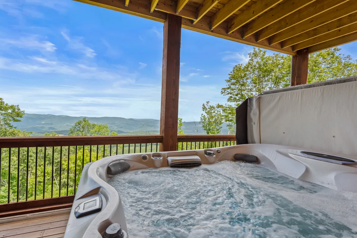 A covered deck area featuring a bubbling hot tub with a mountain backdrop, framed by a sturdy deck railing. The space exudes tranquility, inviting relaxation after a day of adventure while soaking in the views.
