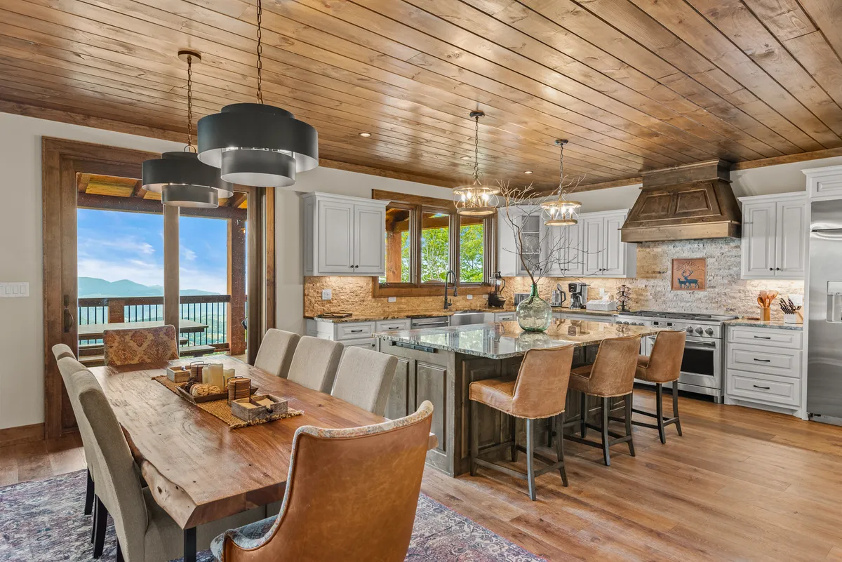 A large wooden dining table with seating for eight is set beside wide windows, with an adjacent kitchen island and modern lighting fixtures overhead. The layout encourages family meals and gatherings, with daylight pouring in and views beyond the glass.