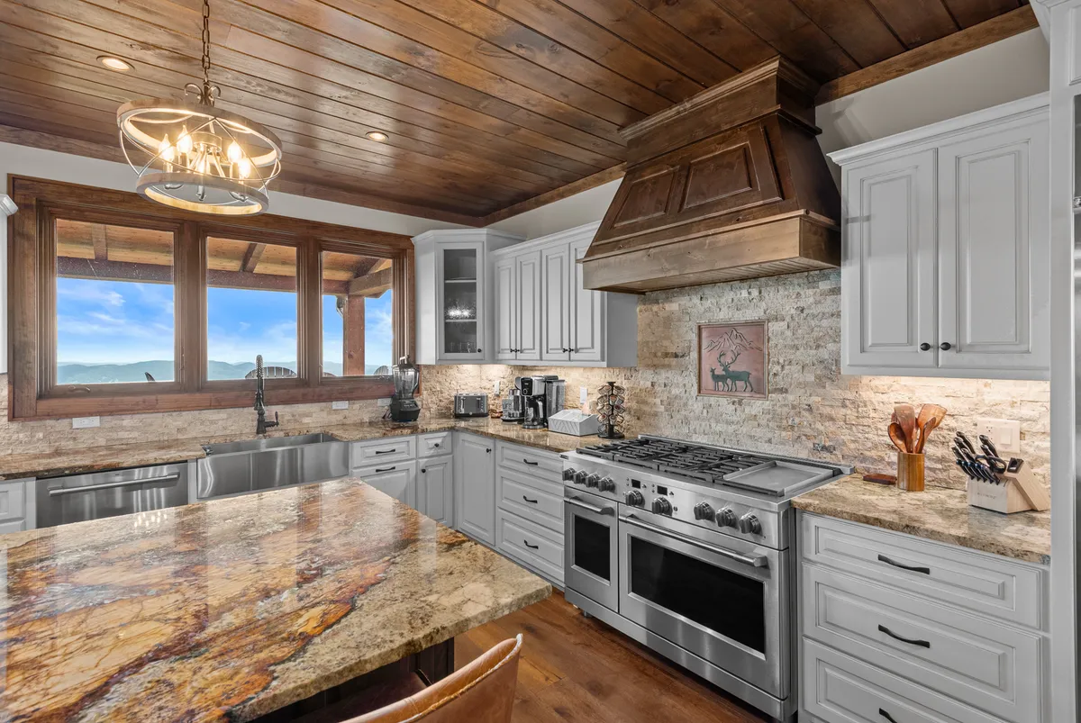 A spacious kitchen with a large granite island, stainless steel appliances, wooden ceiling beams, and panoramic windows that flood the room with light. The scene shows the kitchen as a social hub, ideal for cooking and entertaining, with beautiful views visible.