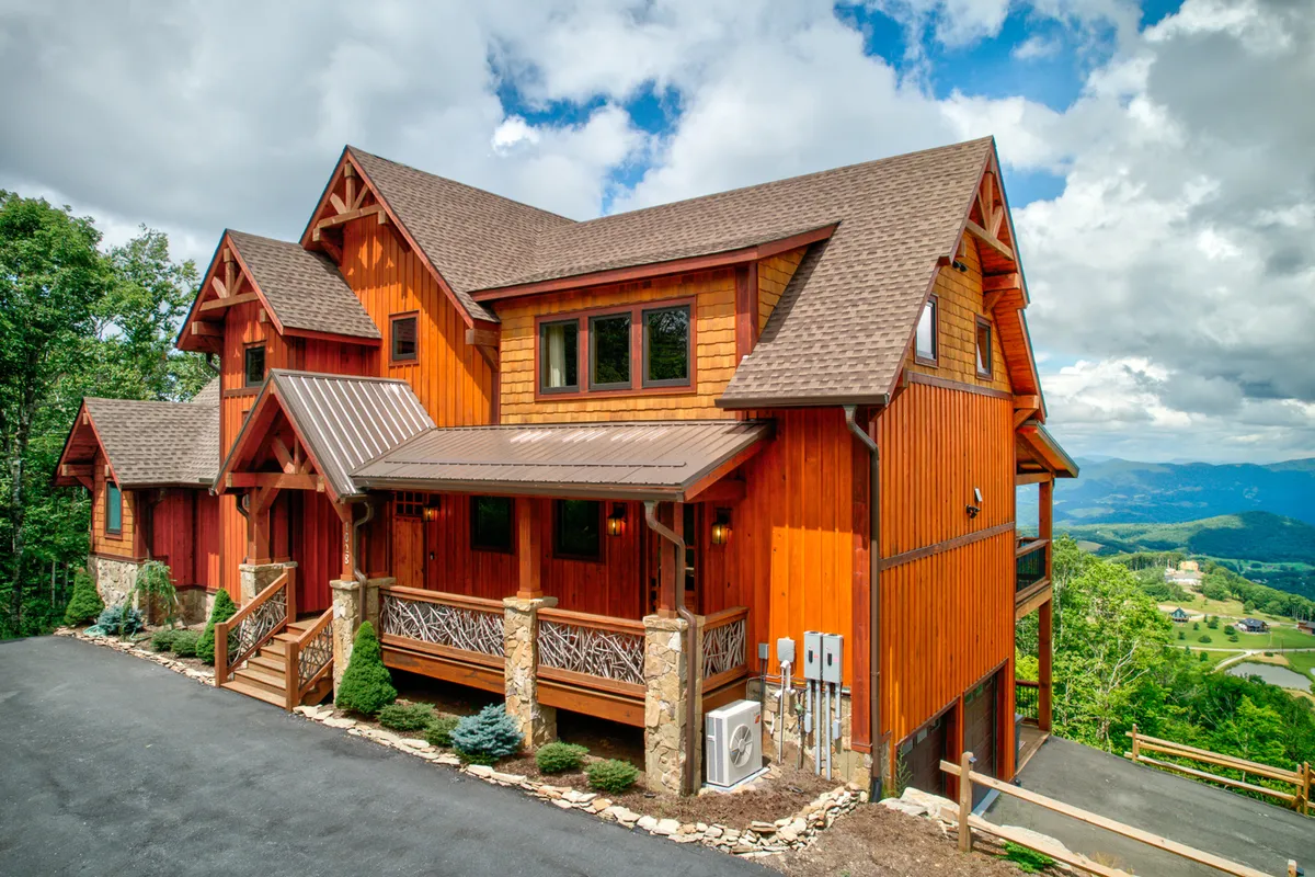 A warmly lit, wooden facade with stone columns and generous windows, surrounded by well-kept landscaping. The wide porch invites guests to relax, socialize, or stargaze while enjoying the fresh mountain atmosphere.