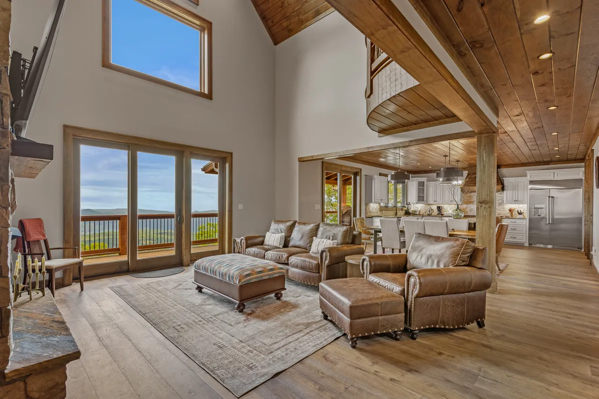 This image features a bright, open-concept living space with large windows offering scenic views, a comfortable seating area, a rustic stone fireplace, and a kitchen layout visible in the background. The room is bathed in natural light, emphasizing its airy and inviting atmosphere.