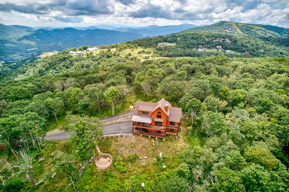The exterior of a large wooden house nestled among trees, with a distinctive circular patio area. The surrounding landscape is peaceful and inviting, hinting at outdoor gatherings, evening bonfires, and quiet moments in nature. The architecture and setting evoke privacy and exclusivity.