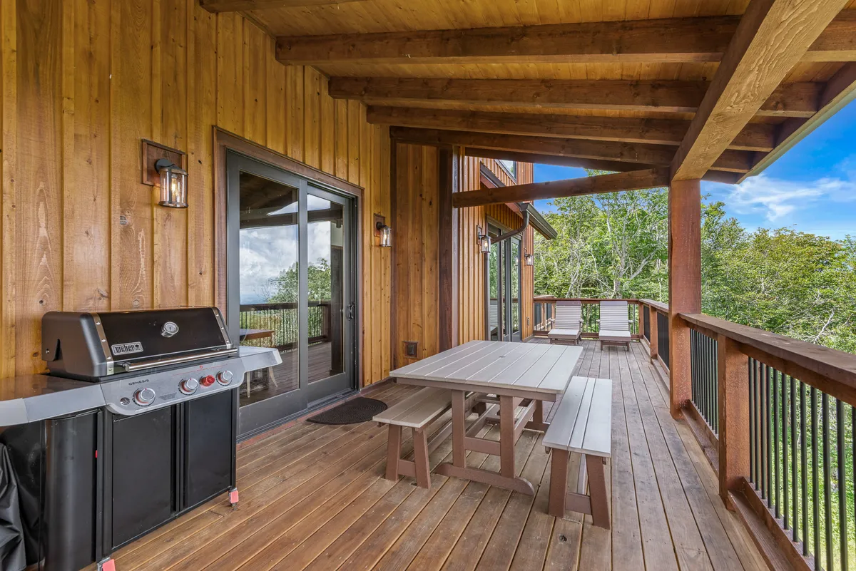 A wooden deck equipped with a large dining table, grill, and sturdy deck railing is framed by surrounding greenery. The setup encourages outdoor dining, barbecues, and socializing, providing a comfortable space for group meals and shared moments against a tranquil mountain backdrop.