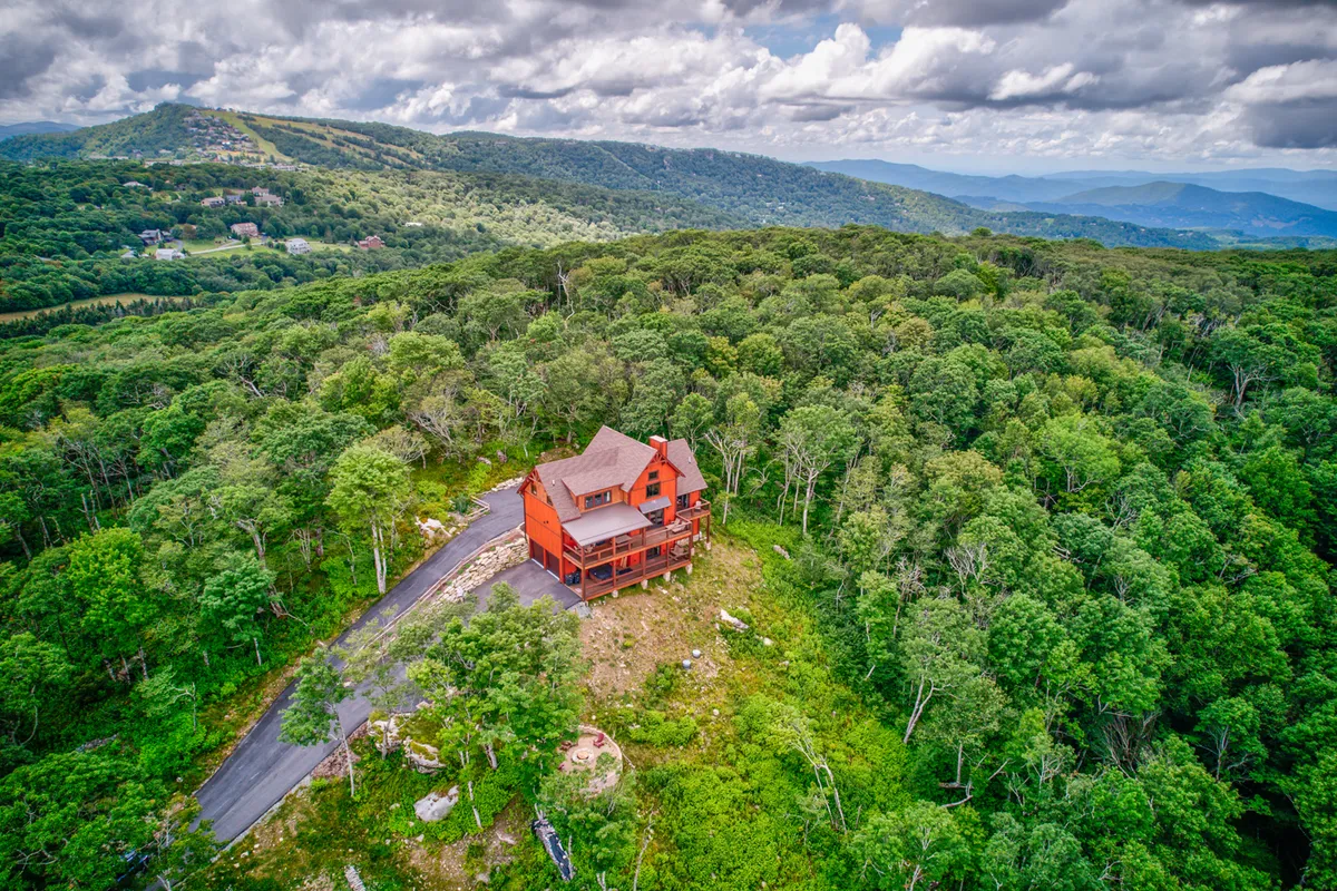 An inviting view of the house nestled among tall trees, with a sweeping mountainous backdrop. The deck offers a prime spot for gathering and taking in the scenery, while the peaceful forest suggests proximity to trails and nature walks.