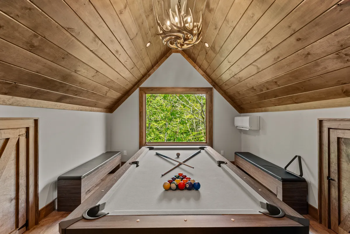 The image features a spacious game room with a pool table positioned under a chandelier, natural light streaming in through a large window, and a cozy wooden ceiling overhead. The setup hints at a multi-purpose area perfect for game nights or casual gatherings.