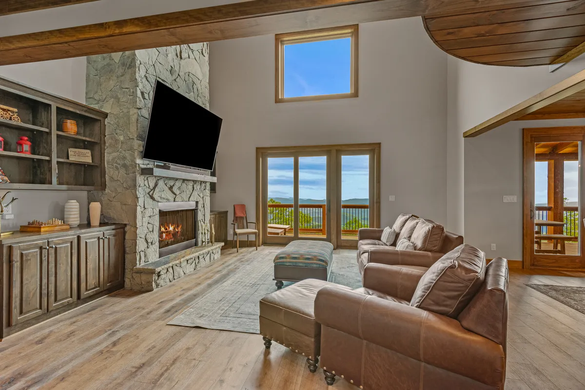 A cozy interior room with a striking stone fireplace, large windows overlooking scenic mountain views, comfortable seating, and wooden shelves filled with books and games. The space radiates warmth and is clearly designed for socializing, relaxing, and enjoying family activities like game nights.