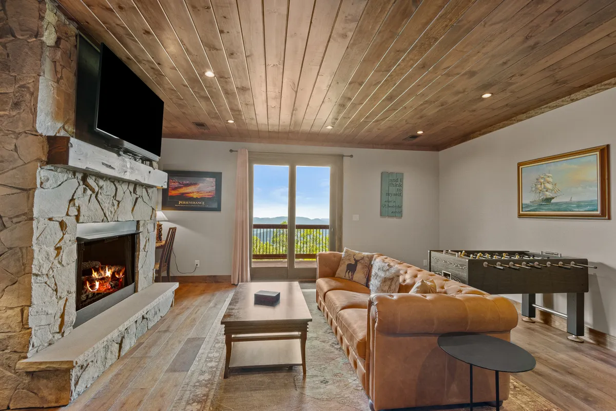 A warm and inviting lower living area with a stone fireplace, leather sofa, large windows with sunset views, and a foosball table. The area is set up for relaxing, playing games, and socializing, making it ideal for family fun or friendly competition.