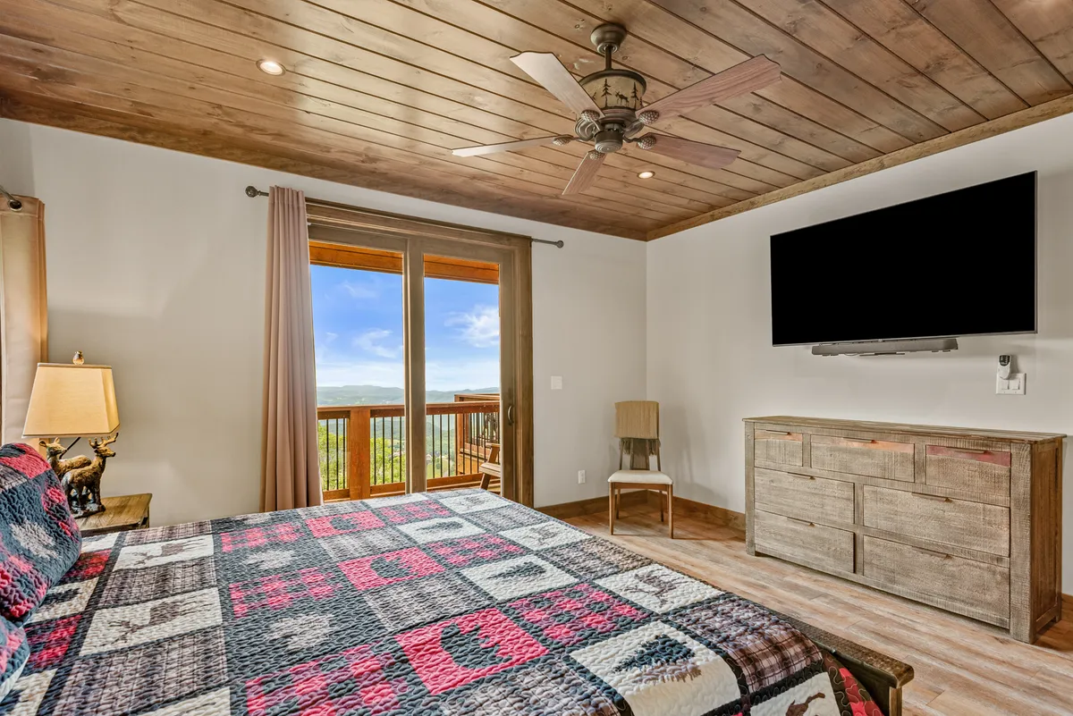 A tranquil king bedroom featuring a bed with a patterned quilt, large windows that frame a stunning mountain view, a wooden ceiling, and a mounted TV. Natural light fills the room, enhancing the sense of spaciousness and retreat.