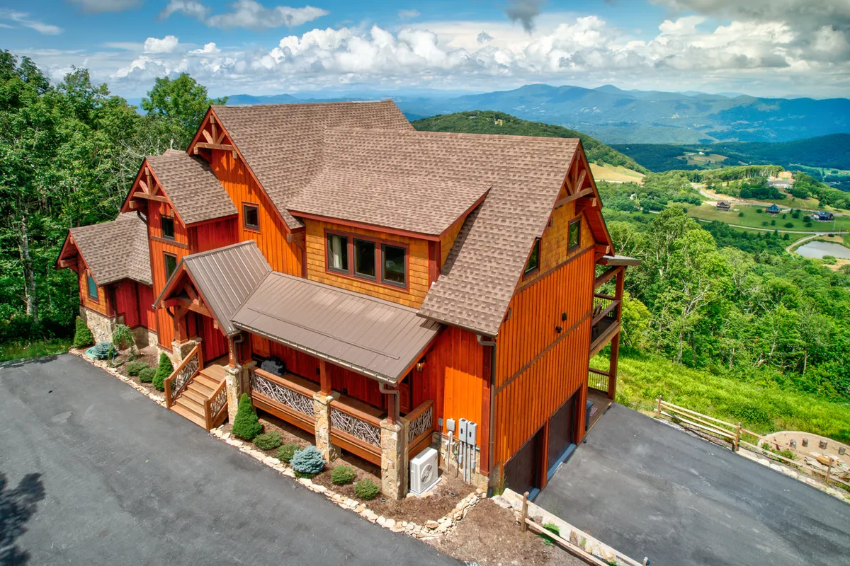 The hero image showcases a beautifully crafted wooden cabin nestled in the mountains of Banner Elk, North Carolina. The cabin stands out with its inviting architecture, surrounded by a landscaped garden and set against a breathtaking mountain backdrop. The spacious porch faces the scenic views, perfect for sipping morning coffee, watching sunsets, or stargazing at night. The driveway welcomes guests into a tranquil, secure environment, ideal for both relaxation and outdoor gatherings.