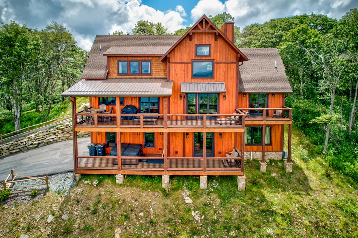 The image shows the wooden exterior of the mountain retreat, featuring multiple balconies and large windows. Lush landscaping surrounds the home, and the scene is bathed in warm, welcoming light—suggestive of an inviting atmosphere ideal for families. The balconies offer space for outdoor relaxation and group activities, with the wooded landscape emphasizing privacy and connection to nature.